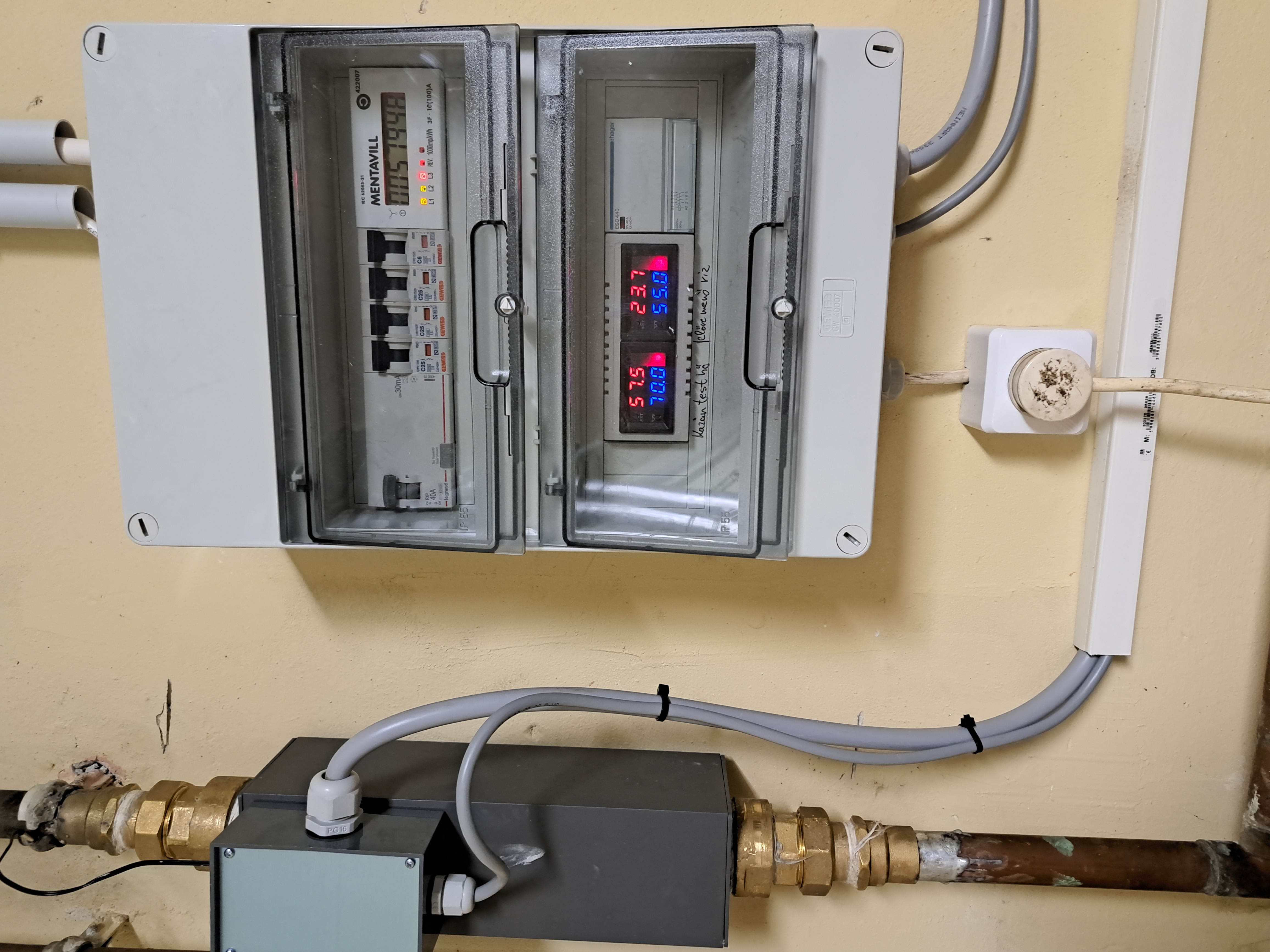 Az AT fűtőmodul egy meglévő rendszerbe kötve: a szürke modul a régi rézcsőbe illesztve, fölötte az elektromos szekrény a fogyasztásmérővel és a hőmérséklet-kijelzőkkel.